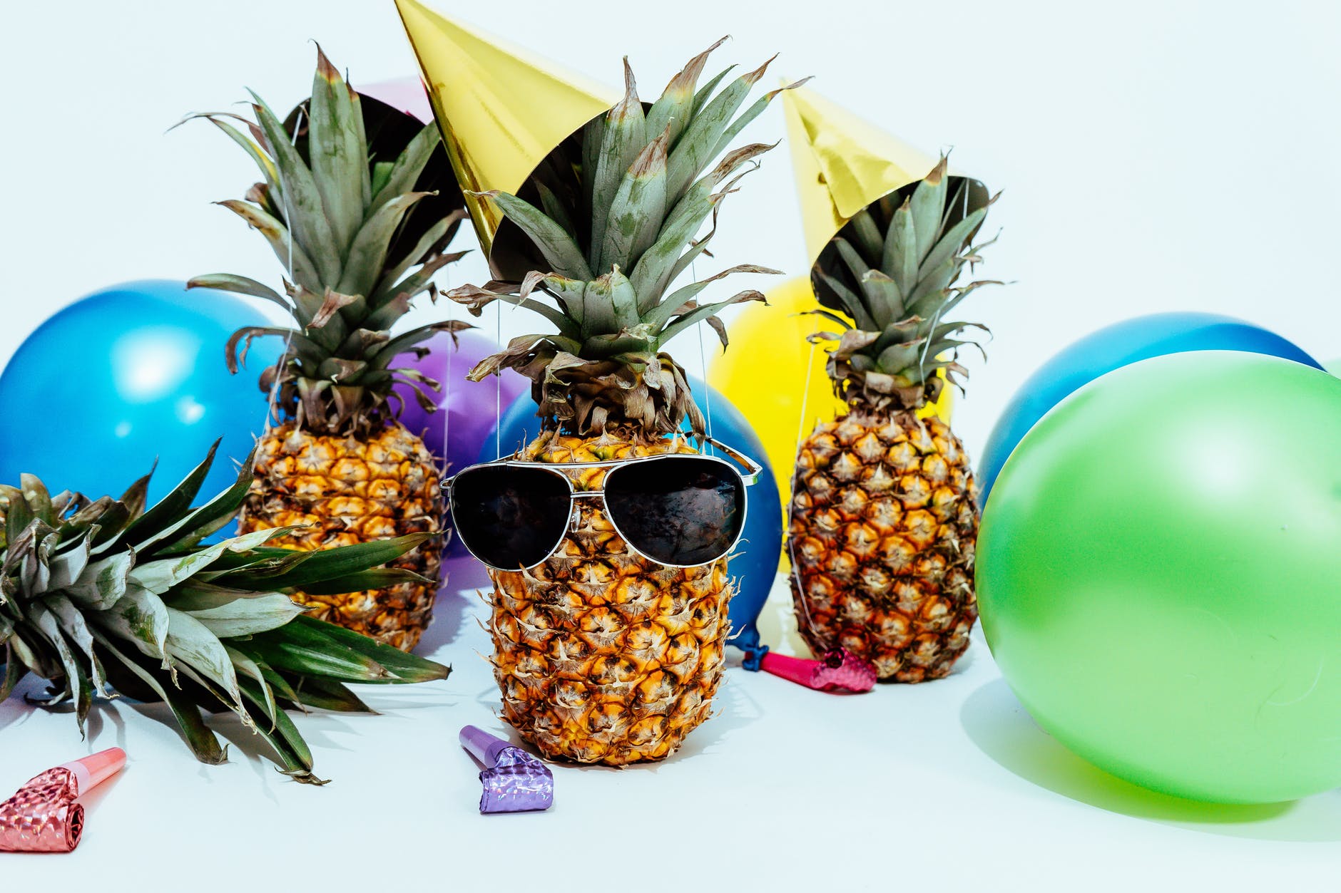 A pineapple wearing sunglasses surrounded by colorful balloons and party hats.