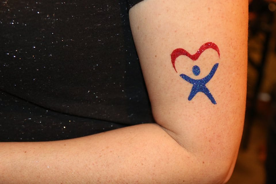 A person's arm with a temporary tattoo of a blue stick figure holding a red heart.