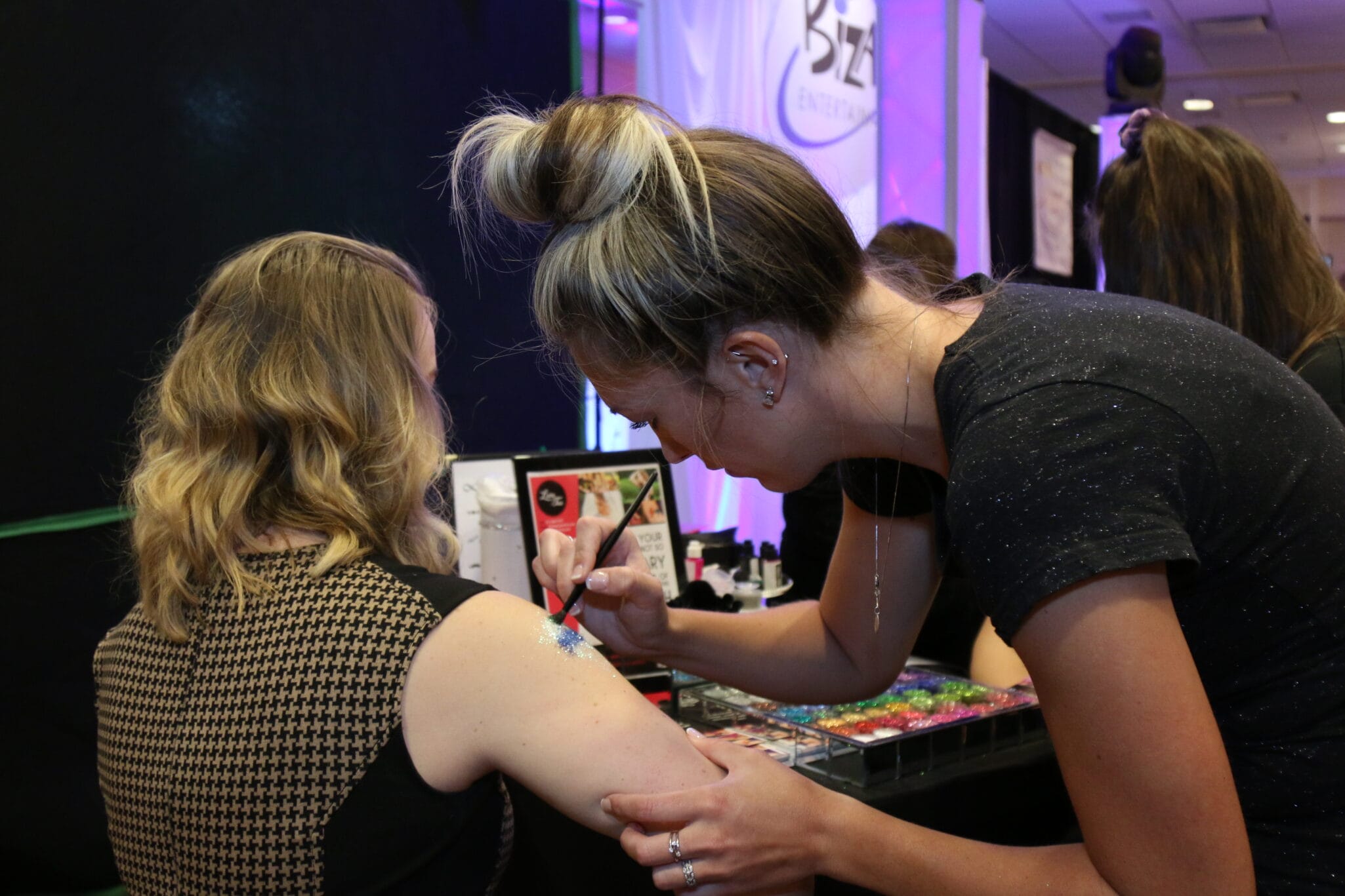 An artist applying body glitter to a woman's shoulder at an event.