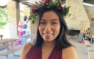 A smiling woman wearing a floral headpiece and a burgundy bridesmaid dress at an outdoor wedding with balloons in the background.